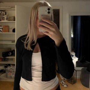 Svart croppad kofta med knappar - Säljer en svart croppad kofta med långa ärmar och knäppning framtill. Enkel och stilren modell som är perfekt att slänga över ett linne eller t-shirt. Koftan har en tight passform och är superenkel att matcha med olika outfits.