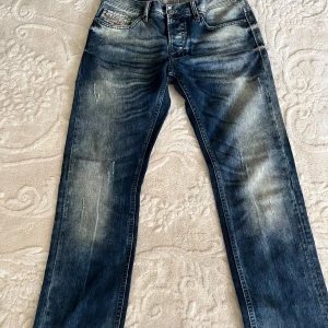 Diesel jeans i tvättad blå denim - Snygga jeans från Diesel i tvättad blå denim med slitningar och ljusa partier. Klassisk femficksmodell med logotypdetalj på myntfickan och tryck på insidan av linningen. Raka ben och normal passform, perfekta till sneakers eller boots.