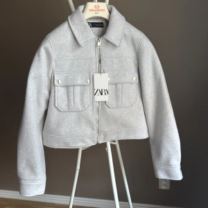 Ljusgrå overshirt från Zara - Snygg ljusgrå overshirt från Zara med klassisk krage, två bröstfickor med knappar och dragkedja framtill. Jackan har en rak, något croppad passform och är tillverkad i ett mjukt ullblandat material. Perfekt för lager-på-lager-stil.