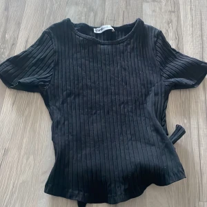 Svart ribbad topp med knyt från FB Sister - Svart ribbad topp från FB Sister i storlek XS. Toppen har korta ärmar, rund halsringning och ett snyggt knytband i midjan som ger en cool detalj. Materialet är mjukt och stretchigt, perfekt för en trendig look.