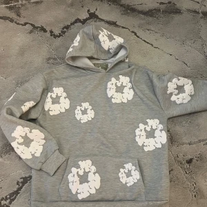 Grå hoodie från Denim Tears med tryck - Grå hoodie från Denim Tears med vita blomliknande tryck över hela tröjan. Tröjan har huva och en klassisk känguruficka framtill. Mjuk insida och avslappnad passform, perfekt för en chill och trendig look.