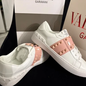 Valentino Rockstud sneakers vit/rosa - Säljer ett par Valentino Rockstud sneakers i vitt skinn med ljusrosa band och ikoniska nitar. Skorna har snörning, platt sula och coola detaljer med pyramidformade nitar både på sidan och sulan. Perfekta för dig som vill sticka ut med en lyxig touch.