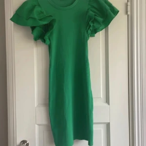 Grön volangklänning från Zara - Supersnygg grön kortklänning från Zara med stora volangärmar som verkligen sticker ut. Klänningen är ribbad och har en tight passform. Perfekt för dig som vill ha något färgstarkt och trendigt i garderoben.