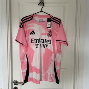 Real Madrid rosa fotbollströja Adidas L - Snygg Real Madrid fotbollströja från Adidas i slim fit-modell. Tröjan är rosa med vita och svarta detaljer, har korta ärmar och svart logga samt klubbmärke på bröstet. Tillverkad i lätt och ventilerande material, perfekt för träning eller match.
