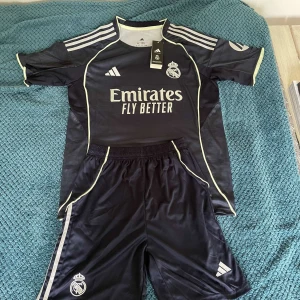 Real Madrid mörkblå matchtröja Adidas M - Säljer en mörkblå Real Madrid fotbollströja från Adidas med korta ärmar och tunna ljusa detaljer. Tröjan har klubbmärke och sponsortryck framtill samt klassiska Adidas-ränder på axlarna. Materialet är lätt och andas, perfekt för träning eller match.