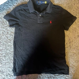 Svart Polo Ralph Lauren Pike - Svart t-shirt från Polo Ralph Lauren med klassisk krage och två knappar framtill. Slim fit-modell med röd broderad logga på bröstet. Tillverkad i mjuk bomull som känns skön mot huden. Perfekt för dig som gillar stilrena och tidlösa plagg. Nypris 850