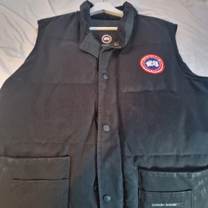 Svart dunväst från Canada Goose - Svart dunväst från Canada Goose med klassisk logga på bröstet. Västen har två stora fickor framtill, tryckknappar och hög krage. Materialet är slitstark polyester och dunfyllning som håller värmen. Perfekt för kalla dagar och snygg att styla över hoodie eller tröja.