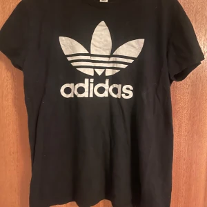 Svart Adidas t-shirt med logga - Svart t-shirt från Adidas med stor vit Trefoil-logga och text på bröstet. Klassisk rund hals och korta ärmar. Perfekt för en sportig och avslappnad stil.