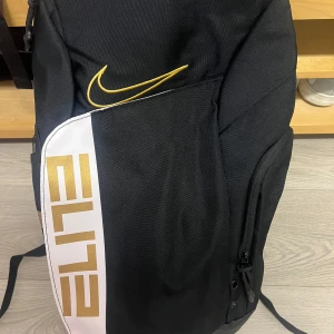 Nike Elite svart ryggsäck - Nike Elite ryggsäck i svart med vita och guldfärgade detaljer. Snyggt broderad Swoosh-logga och ELITE-tryck i guld. Axelremmarna har vadderade paneler och extra komfort. Praktiska fack och dragkedjor, perfekt för träning eller skola.