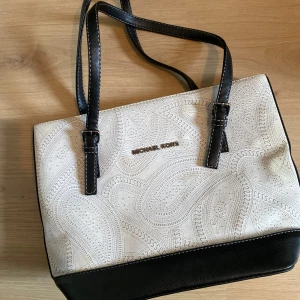 Michael Kors vit och svart handväska - Snygg handväska från Michael Kors i vit och svart med paisley-inspirerat mönster. Väskan har svarta handtag med vita sömmar, silverfärgad dragkedja och flera fack inuti. Materialet är en mix av skinn och textil, perfekt för dig som gillar stilrena detaljer.