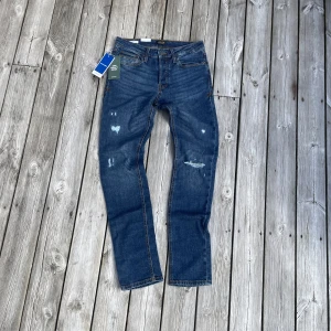 Jack & Jones Slim Straight jeans blå - Snygga blå jeans från Jack & Jones i modellen Slim Straight. Jeansen har klassisk femficksdesign, slitna detaljer framtill och normalhög midja. Materialet är mjuk bomullsdenim med lite stretch för extra komfort. Perfekta för en avslappnad och trendig look. Nypris 800kr!