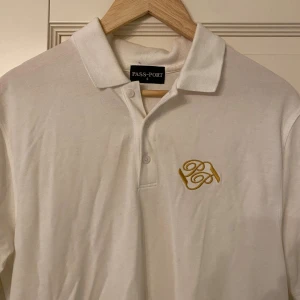 Vit pikétröja från Pass~Port - Vit pikétröja från Pass~Port i 100% bomull med klassisk krage och knäppning framtill. Broderad gul logga på bröstet ger en clean och stilren look. Perfekt för dig som gillar enkel men snygg streetwear.