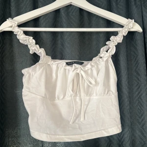 Vit croppad linnetopp från SHEIN - Säljer en söt vit croppad linnetopp från SHEIN med volangiga axelband och knyt framtill. Toppen är ärmlös och har en lätt rynkad design upptill.