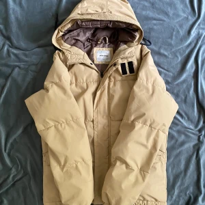 Beige pufferjacka från Jack & Jones - Säljer en beige pufferjacka från Jack & Jones med huva och dragkedja. Jackan har en enkel, clean look med diskreta detaljer och är vadderad för extra värme. Perfekt för kyliga dagar och har en avslappnad passform.