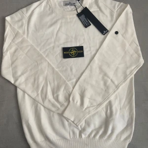 Vit stickad tröja från Stone Island - Vit stickad tröja från Stone Island med klassisk rund halsringning och ikonisk logotyp-patch på ärmen. Tröjan har ribbade muddar och är långärmad, perfekt för lager-på-lager. Snygg och stilren design som passar till det mesta.