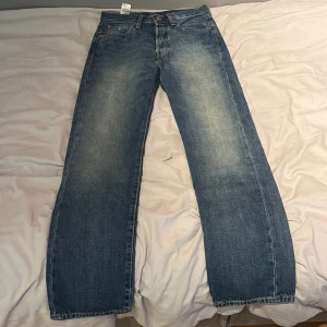 Levi's 501 Premium blå jeans W29 L32 - Klassiska Levi's 501 Premium jeans i mellanblå tvätt med snyggt slitna detaljer. Rak passform och hög midja, tillverkade i robust denim. Perfekta för dig som gillar tidlös stil och vill ha ett par jeans som funkar till allt.