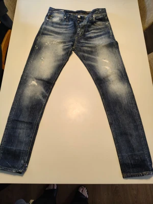 Blå Erik Anti Fit jeans med slitningar - Säljer ett par blå Erik Anti Fit jeans med coola slitningar och tvättade detaljer. Jeansen har klassisk femficksdesign, gul kontrastsöm och en relaxed, loose passform. Materialet är robust denim i bomull och färgen är en snygg mellanblå ton med ljusare partier.