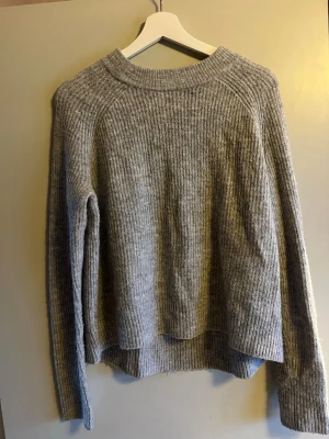 Grå ribbad stickad tröja - Mysig grå stickad tröja med ribbad struktur och rund hals. Tröjan har lång ärm och en något lös passform, perfekt för lager på lager under kyliga dagar. Enkel och stilren design som passar till det mesta.