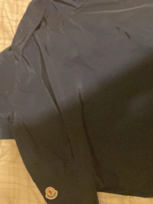 Blå vindjacka från Moncler - Säljer en stilren blå vindjacka från Moncler med klassisk logga på ärmen. Jackan har dragkedja framtill och är tillverkad i lätt polyester, perfekt för vår och höst. Enkel design med lång ärm och diskreta detaljer.