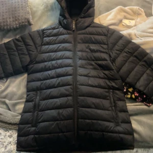 Svart dunjacka med huva - Säljer en svart dunjacka från stadium med huva och dragkedja framtill. Jackan har quiltad design och är perfekt för kyliga dagar. Enkel och stilren look som passar till det mesta.