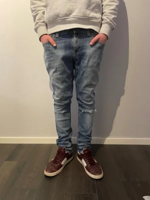 Nudie Jeans Skinny Sam blå jeans - Säljer ett par ljusblå Skinny Sam jeans från Nudie Jeans med slitningar och hål vid knäna. Modellen är skinny fit och har klassiska fem fickor samt broderade detaljer på bakfickorna. Tillverkade i ekologisk bomull och elastan för stretch. Lägg gärna prisförslag!!