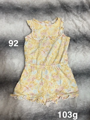 Blommig byxdress från Newbie, strl 92 - Supersöt byxdress från Newbie i ljusgul och pastellblå med blommigt mönster. Volangdetaljer vid axlar och ben, resår i midjan. Perfekt för varma dagar och riktigt gullig stil!🌸Fint använt skick. Storlek 92