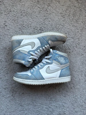 Nike Air Jordan 1 High ”hyper royal” blå/vit - Säljer ett par Nike Air Jordan 1 High sneakers i vitt och ljusblått med grå detaljer. Skorna har klassisk snörning, rund tå och perforerade partier på tån. Ovandelen är i mocka och skinn, med ikoniska Nike-loggor på sidan och tungan. Perfekta för dig som gillar streetwear.