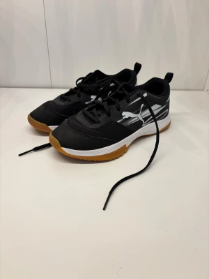 Svarta Puma sneakers strl 38.5 - Säljer ett par svarta Puma sneakers med vita detaljer och gummisula. Skorna har snörning, meshpaneler och logga på sidan. Modellen heter Varion och passar perfekt för dig som gillar sportig stil. Snygg kontrast mellan svart ovandel och vit sula med brun undersida.