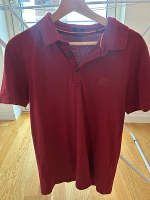 Röd pikétröja från BOSS Slim Fit M - Säljer en röd pikétröja från BOSS i storlek M med slim fit-passform. Tröjan har klassisk krage, två knappar framtill och diskret BOSS-logga på bröstet. Tillverkad i mjuk bomull och har en enkel, stilren look.