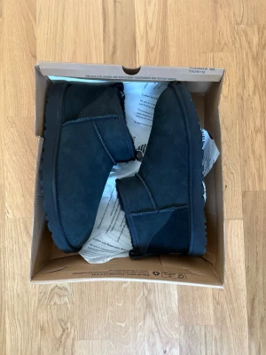 Svarta UGGs - Säljer ett par helt nya klassiska svarta UGG boots i mjuk mocka med rund tå och platt sula. Insidan är fodrad med fluffigt material för extra värme. Skorna har en enkel design med diskret UGG-logga bak på hälen och dragflik för enkel påtagning. Storlek 44 men små i storleken. 