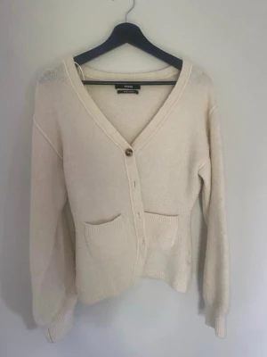 Kofta från BikBok - Mysig beige kofta från BikBok i wool blend. Koftan har v-ringning, knappar framtill och två praktiska fickor. Långärmad modell och en enkel, clean look som passar perfekt till lager på lager.💕den är typ vit/creme vit/beige