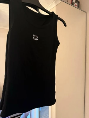 Svart ribbad linnetopp  - Svart ribbad linnetopp från Miu Miu med vit logotyp framtill. Toppen är ärmlös och har en klassisk rund halsringning. Perfekt för en stilren och enkel look, lätt att matcha med olika outfits. Köpt i Thailand 