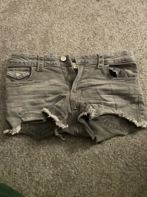 Grå slitna shorts från Gina Tricot - Grå denimshorts från Gina Tricot med råa, fransiga kanter och slitningar. Klassisk femficksmodell med knapp och dragkedja framtill samt bälteshällor. Bakfickor med lock och knappdetalj. Perfekta för en avslappnad look.
