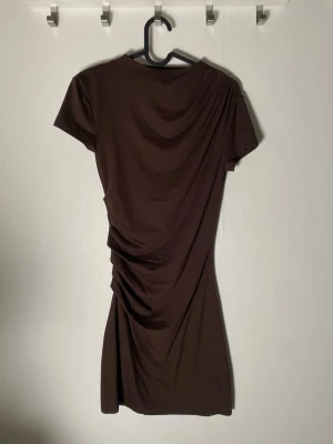 Brun kortklänning från Divided - Säljer en brun kortklänning från Divided i mjuk bomull. Klänningen har korta ärmar och snygga draperade detaljer längs sidan som ger en cool och modern look. Perfekt för dig som gillar minimalistisk stil med en twist.