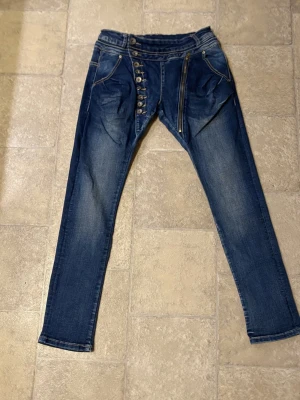 Blå jeans med knappar och dragkedja - Säljer ett par blå jeans med unika detaljer som sned knäppning med flera knappar och en synlig dragkedja framtill. Jeansen har smal passform och klassiska fickor fram och bak. Perfekta för dig som vill sticka ut lite extra!