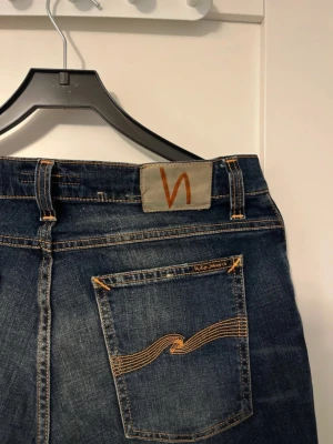 Mörkblå Nudie Jeans 32/32 - Säljer ett par mörkblå jeans från Nudie Jeans med klassisk orange söm och vågmönster på bakfickorna. Modellen har normal passform och raka ben. Tillverkade i slitstarkt jeanstyg med snygga tvättade detaljer. Perfekta för dig som gillar stilrena och tidlösa jeans.