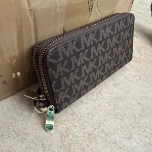 Brun MK plånbok med logomönster - Snygg brun plånbok från Michael Kors med MK-logga över hela ytan. Plånboken har dubbla dragkedjor i guld, flera kortfack och handledsrem. Tillverkad i tåligt syntetmaterial och har ett stilrent, rektangulärt format.