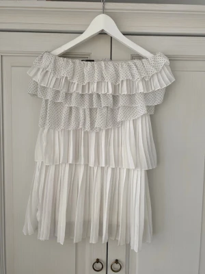 Vit axelbandslös volangklänning - Supersöt axelbandslös kortklänning i vitt med flera lager volanger och små prickar upptill. Klänningen har plisserad kjol och är gjord i ett lätt, luftigt material som känns perfekt för sommaren. Passformen är loose och modellen är riktigt flowy och lekfull. Storlek 38