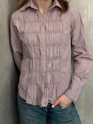 Ljuslila skjorta från Fransa - Ljuslila skjorta från Fransa med långa ärmar och knappar framtill. Skjortan har snygga rynkade detaljer på framsidan som ger en unik look. Tillverkad i mjuk bomull och har klassisk krage. Perfekt att styla med jeans eller kjol.