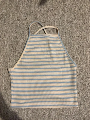 Dam Randig ribbad linnetopp i pastell - Säljer en söt ribbad linnetopp med tunna axelband som korsas i ryggen. Toppen har ljusblå och vita horisontella ränder och är croppad i modellen. Perfekt för varma dagar och enkel att matcha med jeans eller kjol i storlek XS.