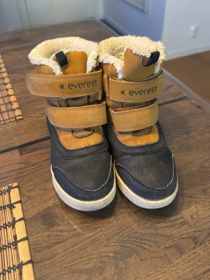 Everest fodrade boots i svart och beige - Fodrade boots från Everest i storlek 27, med svart och beige färg. Skorna har två kardborreband för enkel påtagning och en mjuk, fluffig insida. Yttermaterialet är en mix av syntet och skinn, med robust gummisula som ger bra grepp. Perfekta för kalla dagar. I mycket bra skick. Använda av ett barn.