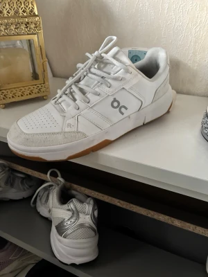 Vita sneakers från DC med gummisula 41  - Säljer ett par fräscha vita sneakers från DC med klassisk design. Skorna har snörning, perforerad tå och detaljer i mocka samt en snygg gummisula i brunt. Märkets logga syns på sidan och baksidan har en liten röd detalj. Perfekta för en clean och sportig look.