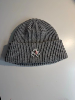 Grå ribbstickad mössa från Moncler - Säljer en snygg grå ribbstickad mössa från Moncler med uppvikt kant och klassisk Moncler-logga framtill. Mössan är tillverkad i mjuk ull och har en clean, stilren look som passar perfekt till kalla dagar.