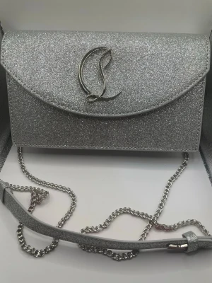 Glittrig silver axelväska från Louboutin - Säljer en glittrig silverfärgad axelväska från Louboutin med kedjerem och magnetlås. Väskan har ett stilrent L-märke på framsidan och insidan är röd med flera fack för kort och småsaker. Perfekt för dig som vill sticka ut med en snygg accessoar.