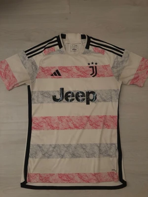Juventus bortatröja Adidas S - Juventus bortatröja från Adidas i storlek S. Tröjan är vit med breda ränder i rött och grått, svart Adidas-logga och Juventus-emblem med tre stjärnor. Materialet är polyester och tröjan har svarta detaljer längs axlar och sidor. Perfekt för fotbollsfans!