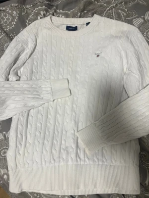 Vit kabelstickad tröja från GANT - Säljer en vit kabelstickad tröja från GANT med rund hals och diskret logga broderad på bröstet. Tröjan har långa ärmar och ribbade muddar vid ärmslut och nederkant. Perfekt för dig som gillar klassisk och stilren design.