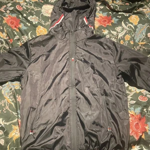 Svart vindjacka från Moncler med huva - Svart vindjacka från Moncler med huva och snygga rödvita detaljer på insidan av huvan. Jackan har dragkedja framtill, två sidofickor med dragkedja och Moncler-logga på ärmen. Materialet är lätt och syntetiskt, perfekt för blåsiga dagar.