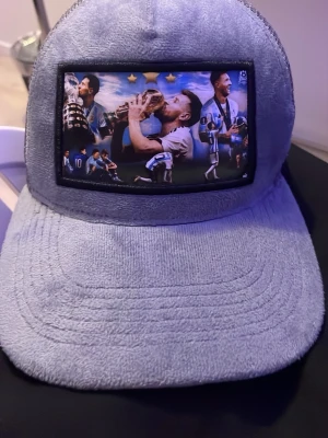 Grå meshkeps med fotbollstryck - Säljer en grå keps med mesh baktill och justerbar knäppning. Framsidan har ett stort tryck med fotbollsmotiv och blå detaljer. Kepsen har en mjuk, frottéliknande känsla framtill och är perfekt för dig som gillar sportig stil.