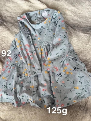 Blommig ljusblå klänning - Supersöt ljusblå klänning med blommigt mönster i rosa, gult och svart. Klänningen har lång ärm, rund hals. Perfekt för dig som gillar färgglada och lekfulla plagg!🌸 Fint använd skick. Storlek 92