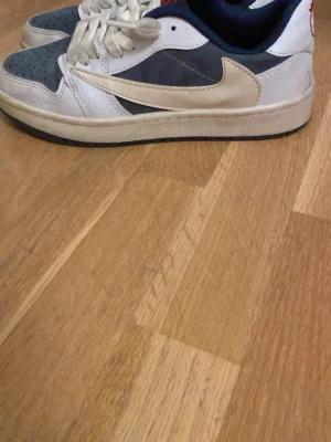 Nike Air Jordan 1 Low blå/vit - Säljer ett par Nike Air Jordan 1 Low med vit och marinblå ovandel, beige swoosh och vita snören. Skorna har klassisk rund tå, platt sula och är tillverkade i skinn och syntet. Perfekta för dig som gillar streetwear och sneakers med unik design.